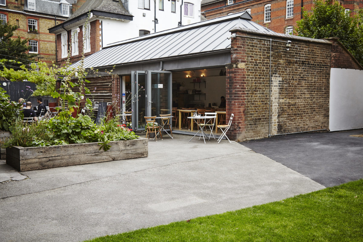 Hand-drawn ink illustration of a small historic brick building with an open shaded patio and surrounding garden greenery, muted earth tones and warm light, inviting calm mood.